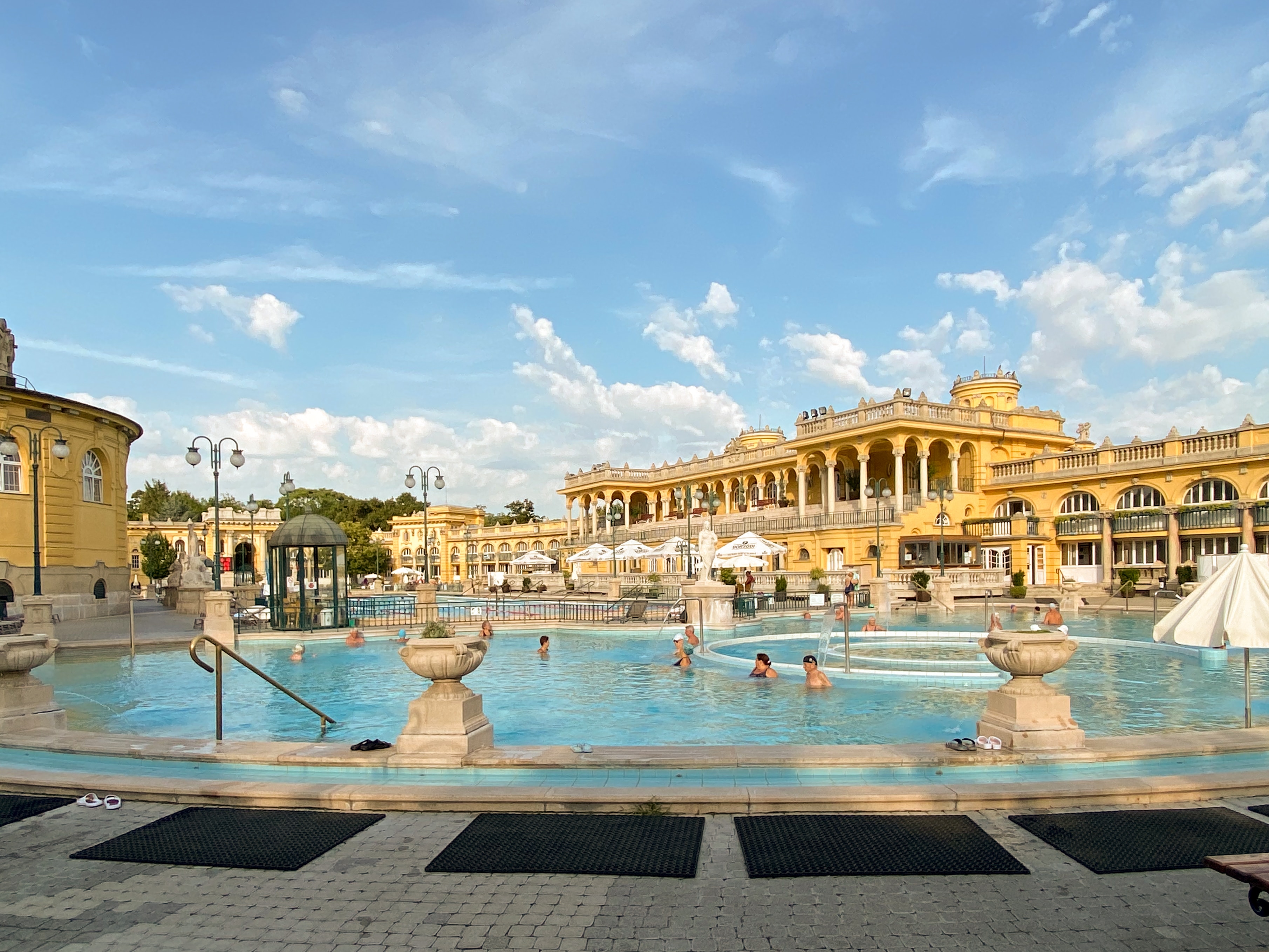 An outdoor thermal bath in EOR Hungary with visitors enjoying the water against the backdrop of a classical architecture building under a blue sky with scattered clouds.